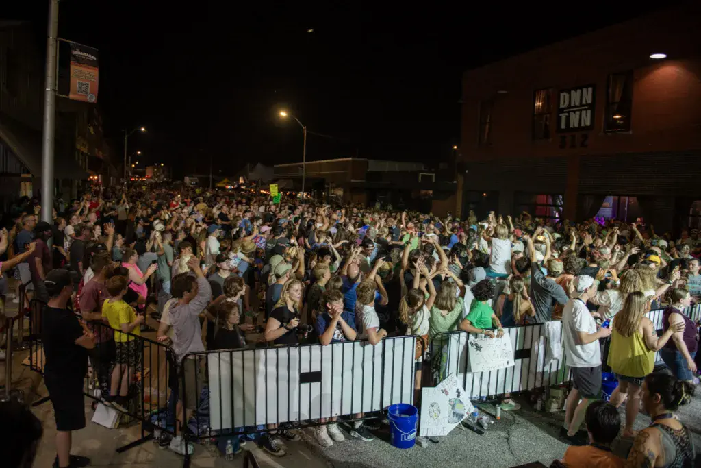 A large crowd of festival attendees with raised hands fills a downtown street at night, separated by white barriers.
