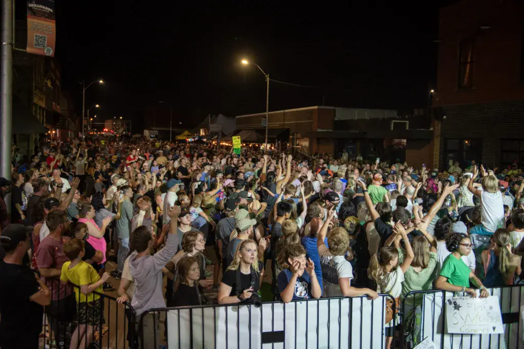 A large crowd of festival attendees dancing and celebrating at night under street lights at an outdoor music festival.