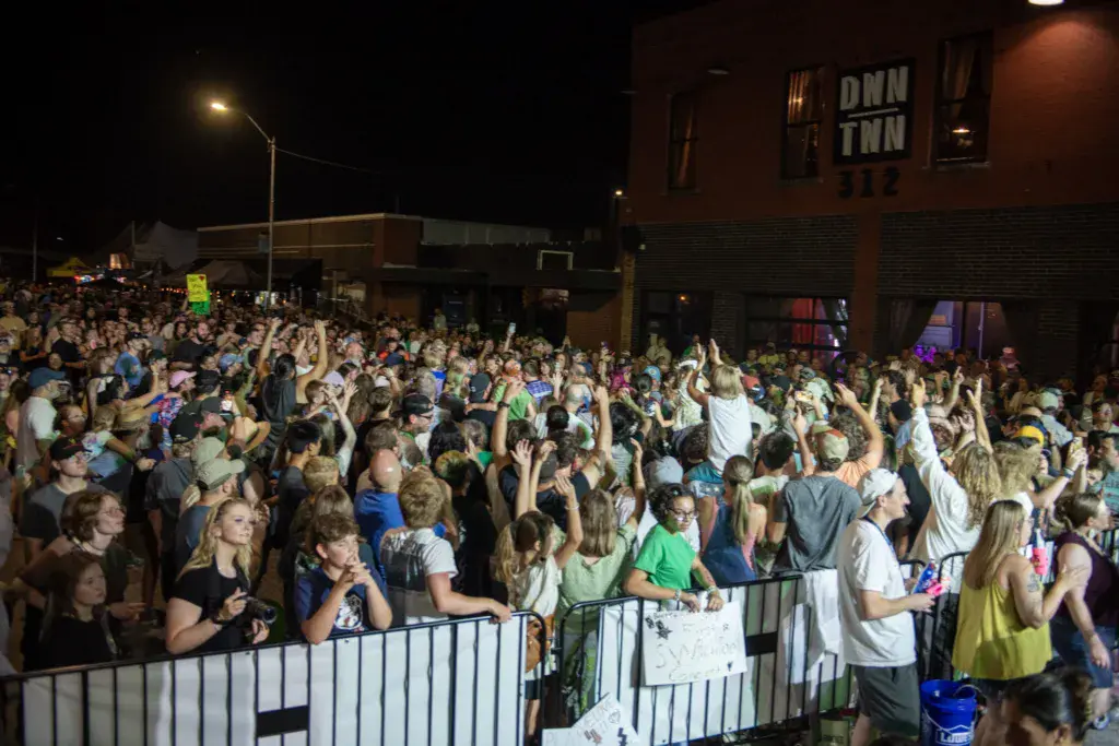 A large crowd gathers at night at Dancing Rabbit Music Festival, with raised hands and barriers visible under street lighting.