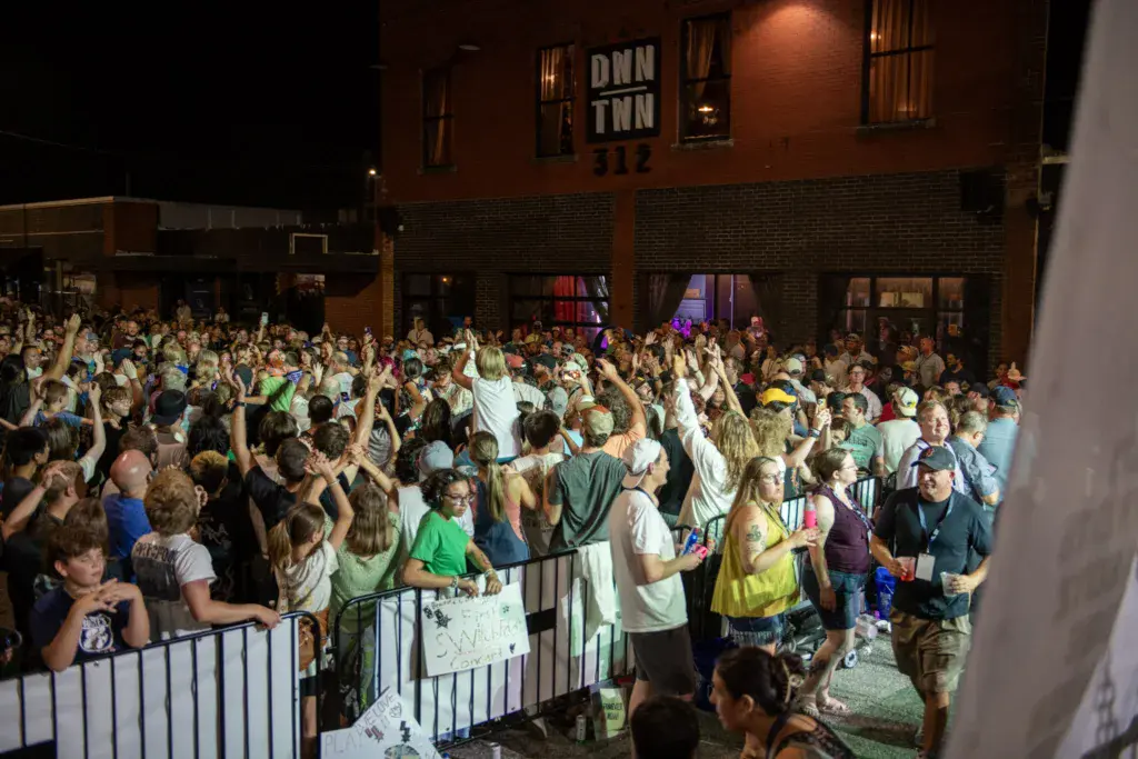 A large crowd of festival attendees with raised arms dancing and celebrating in front of a brick venue building at night.