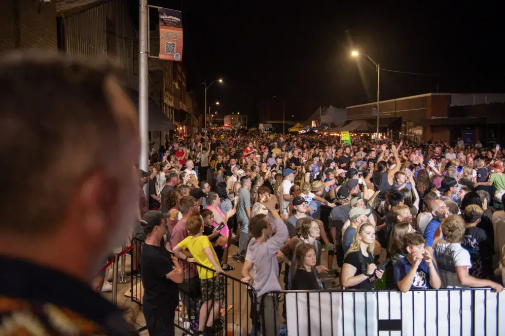 A large crowd of festival attendees gathered at night in an outdoor venue with stage lights and signage visible.