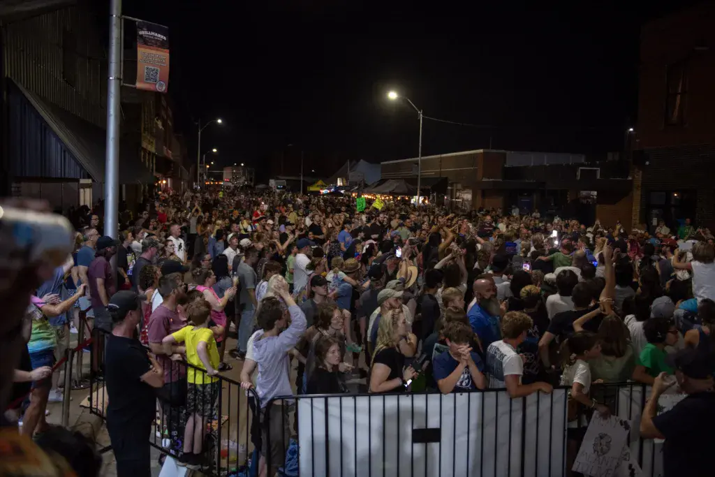A large crowd of festival attendees fills an outdoor venue at night under bright lights.