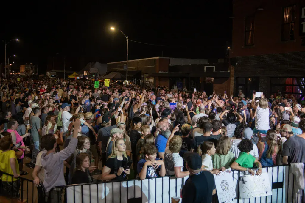 Large crowd of festival attendees gathered at night in an outdoor urban venue, many holding phones and waving hands.