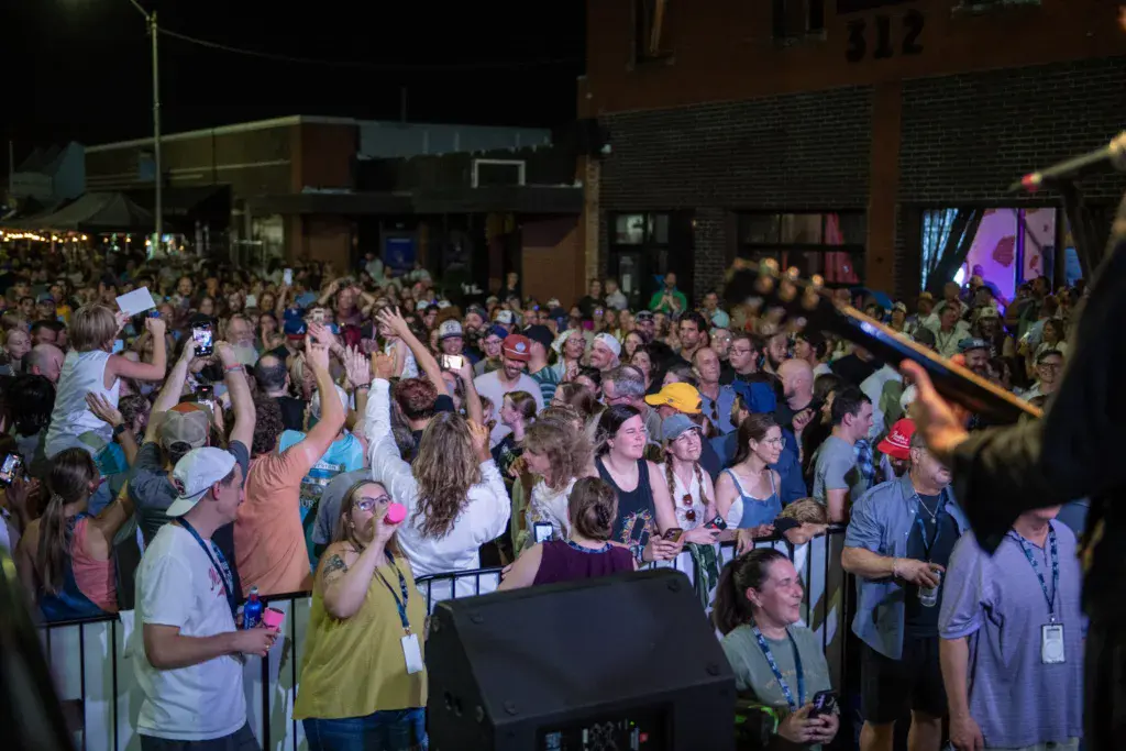 Large outdoor crowd at night watches a performance, many holding up phones and cameras to record.