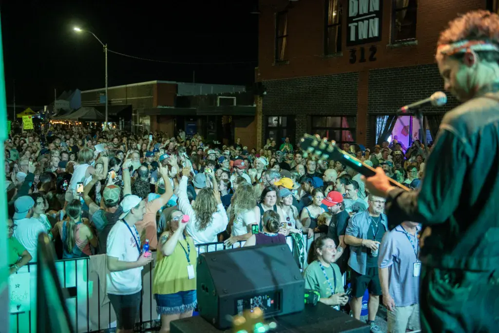 A performer on stage sings to a large outdoor crowd at night, with brick buildings and street lights visible in the background.