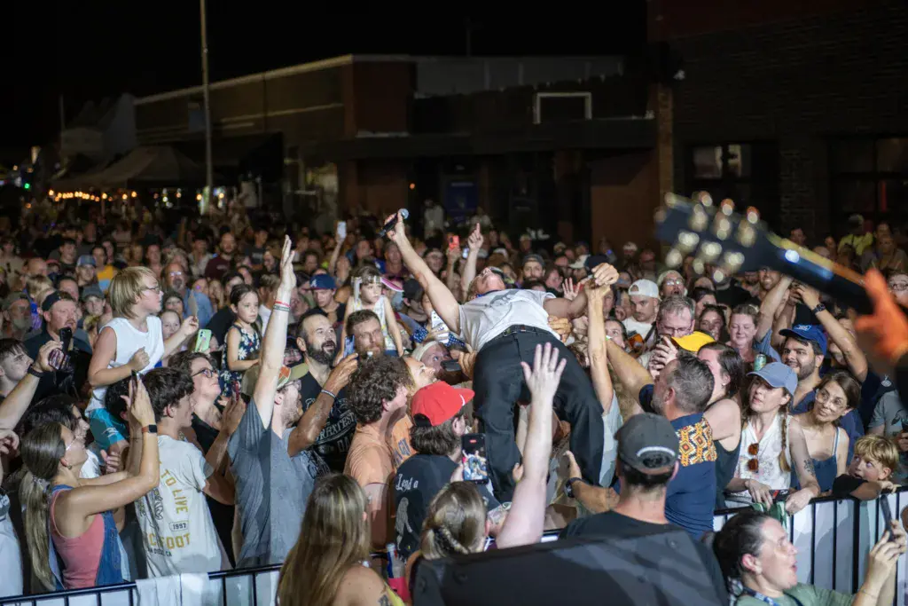 A crowded audience at night lifts a person in the air during a lively music festival performance.