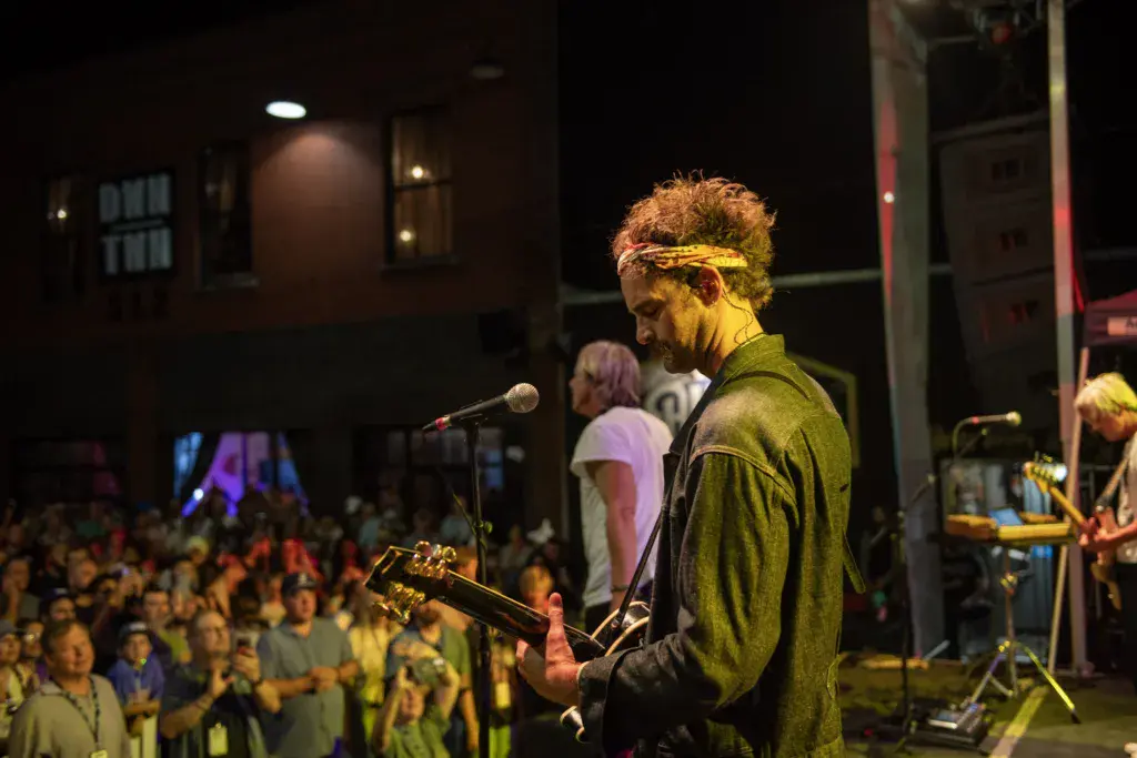 A performer with curly hair and a yellow headband plays guitar at a microphone on an outdoor stage at night, facing a large crowd.