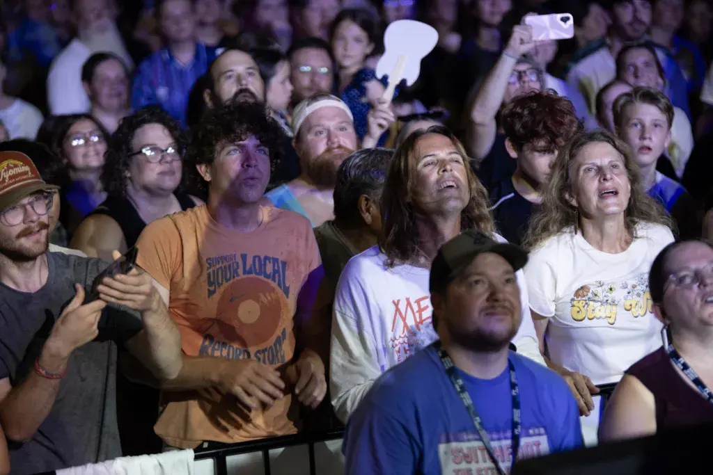 A diverse crowd of festival attendees watches a performance at night, many looking upward with engaged expressions.
