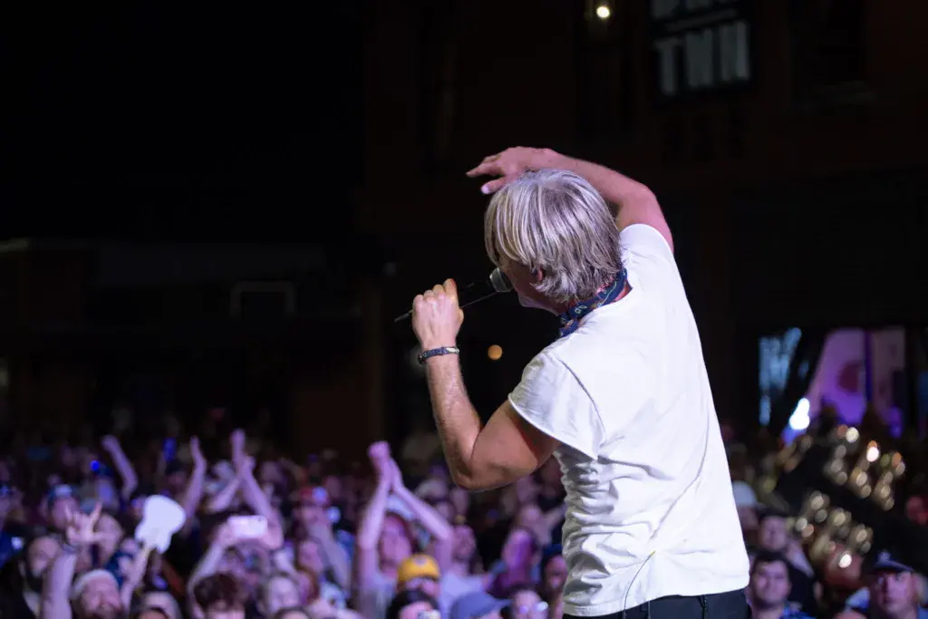 A performer with gray hair in a white shirt sings into a microphone on stage before a large crowd at night.