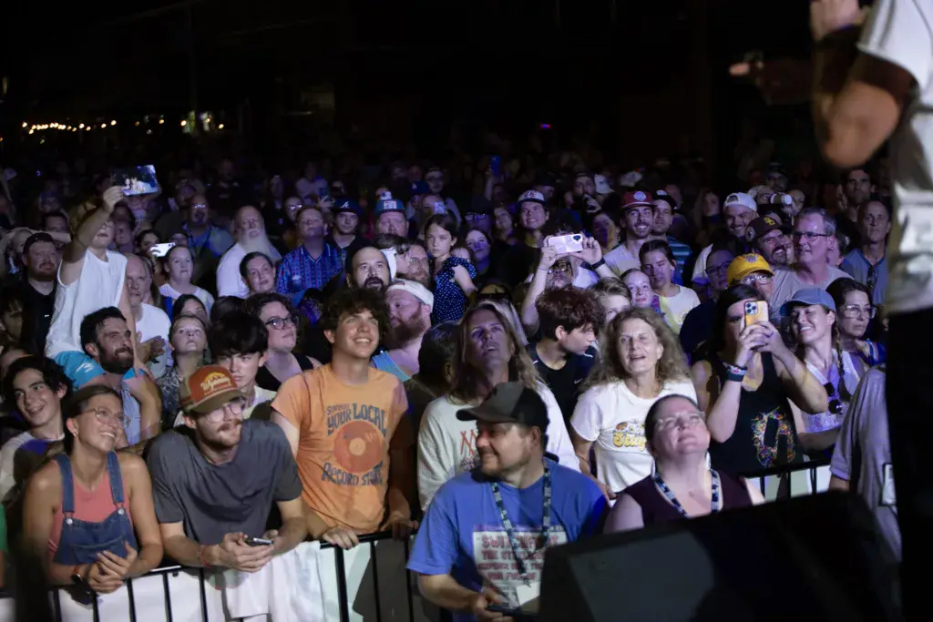 A large crowd of festival attendees watches a performance at night, many looking upward and some holding up phones to record.