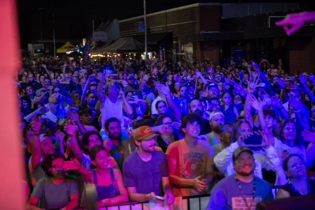 Large crowd of festival attendees dancing and celebrating under vibrant blue and pink stage lighting at night.