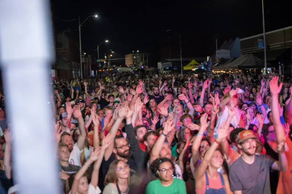Thousands of festival attendees with hands raised under pink stage lighting at night.