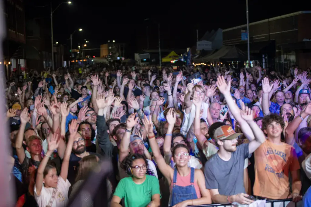 Large crowd at night with hands raised, illuminated by stage lighting at an outdoor music festival venue.