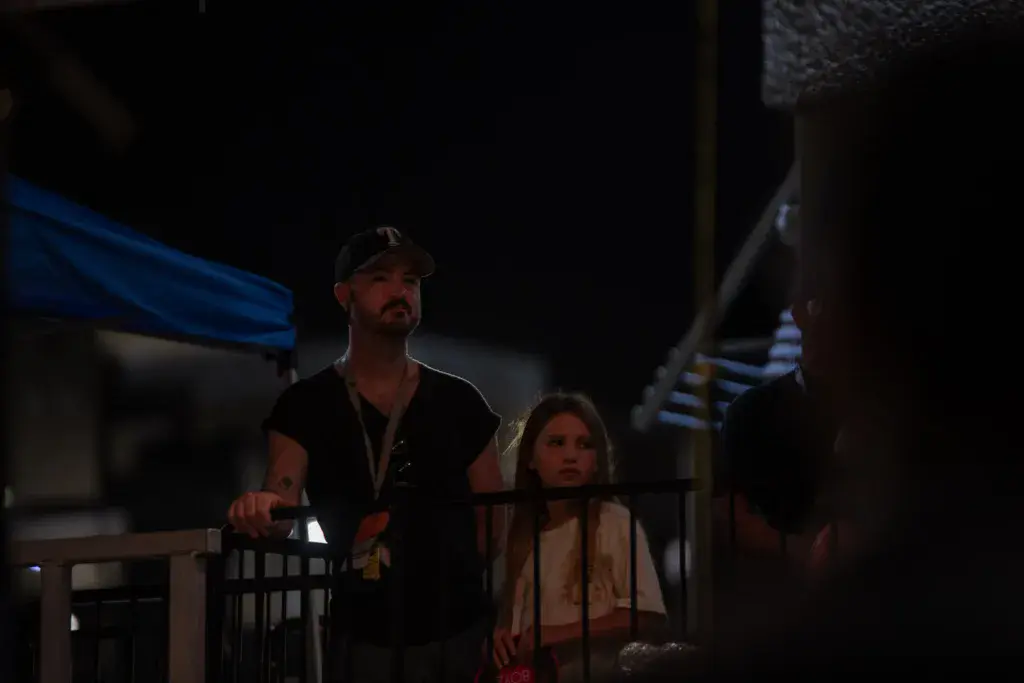Two people stand at a festival railing at night with venue structures and blue lighting visible in the background.