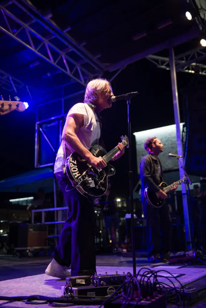 Two musicians perform on a stage bathed in blue lighting, one playing guitar and singing, the other playing bass guitar.