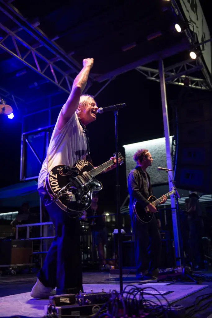 A guitarist raises his fist triumphantly on stage while singing into a microphone, with another band member visible playing guitar beside him.