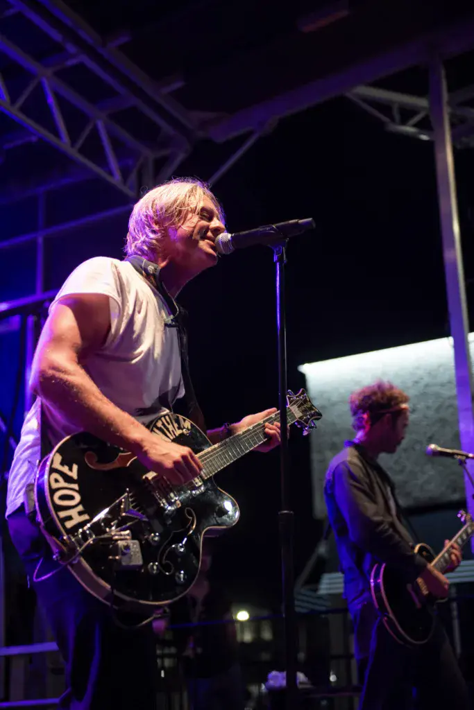 A musician in a white t-shirt sings into a microphone while playing an electric guitar on stage under purple lighting.