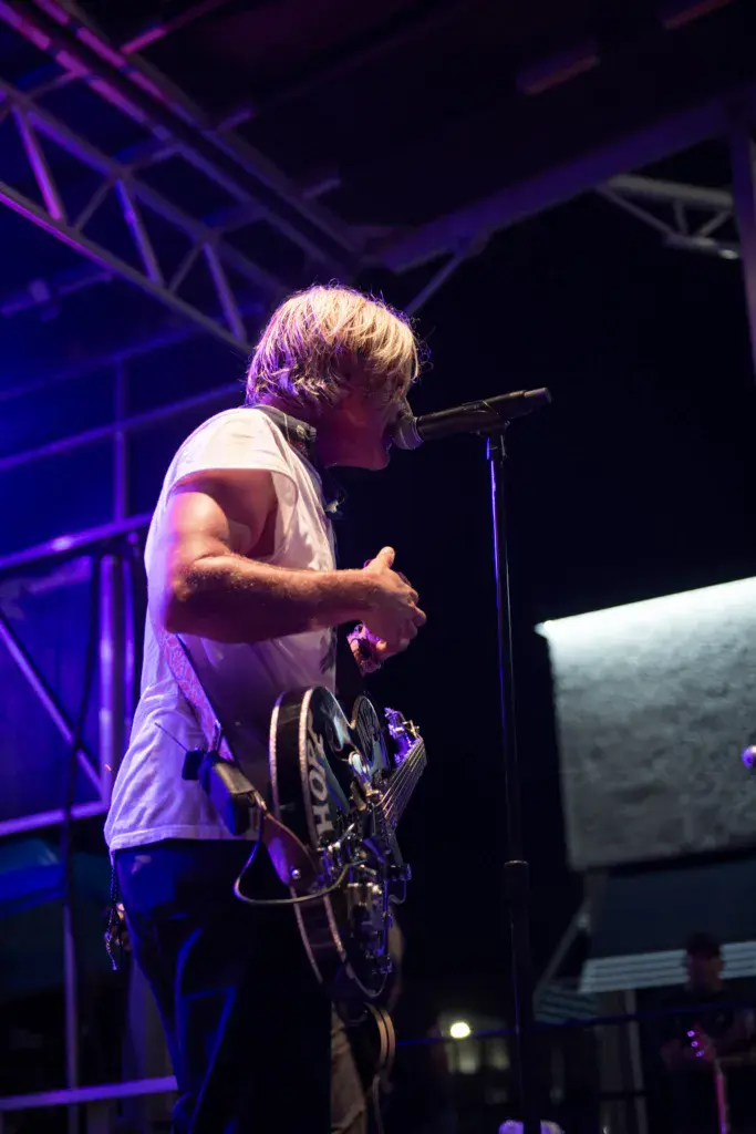 A performer singing into a microphone on stage under purple lighting at an outdoor music festival.