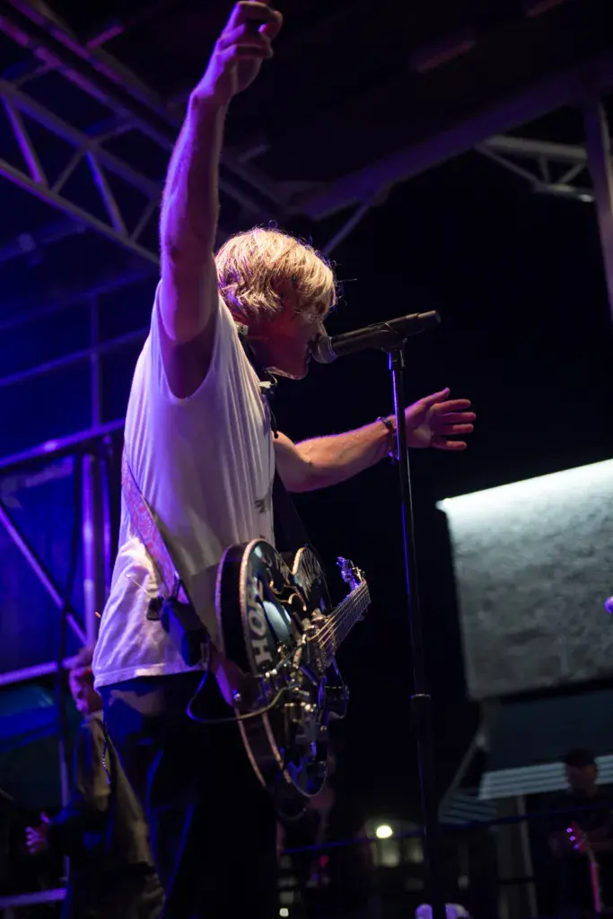 A performer with blonde hair singing at a microphone while raising their fist, illuminated by purple stage lighting and holding a guitar.