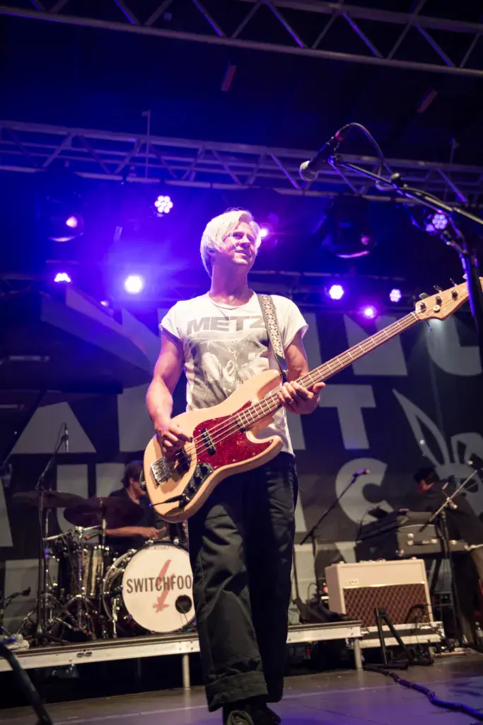 Bassist performing on stage with purple lighting and band name visible on drum kit behind.