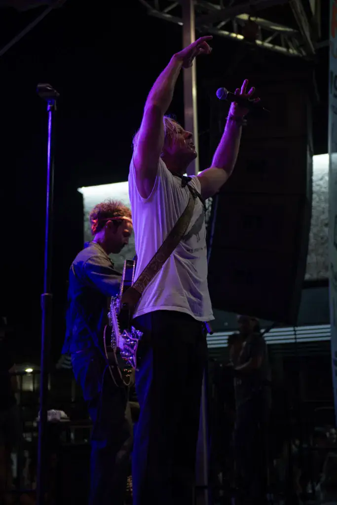 A performer on stage raises both arms in celebration while a saxophonist plays beside them under purple stage lighting.