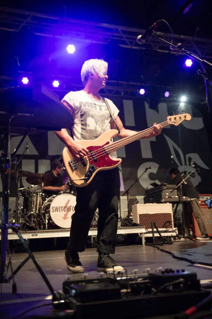 Bass player performing on stage under blue lighting at an outdoor music festival.