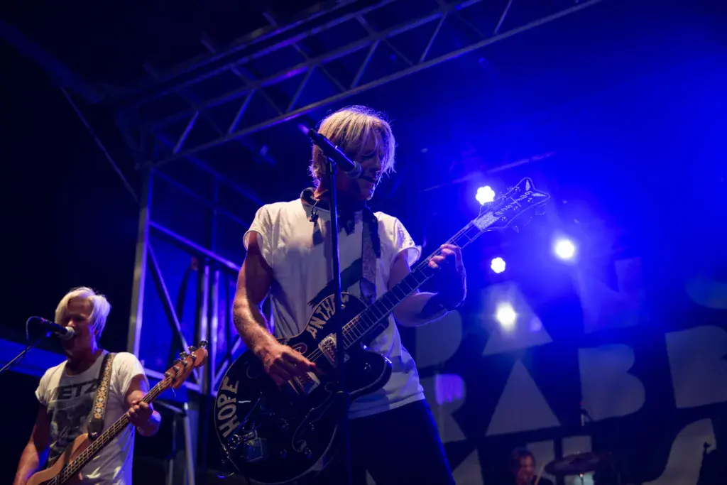Two musicians perform on stage under bright blue stage lighting, with one singing at the microphone and another playing guitar.