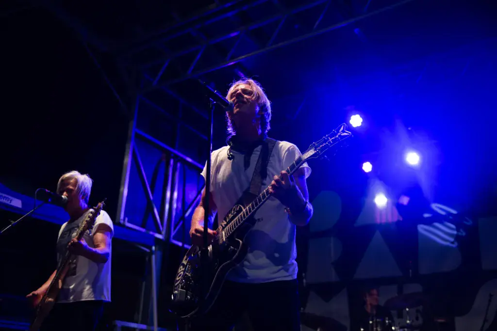 Two musicians perform on stage under blue lighting at an outdoor music festival.