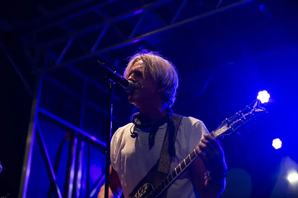 A musician with light-colored hair plays bass guitar on stage under blue lighting.