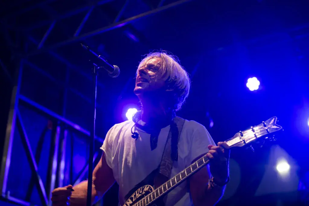 A musician with blonde hair plays bass guitar on stage under blue stage lighting at the festival.