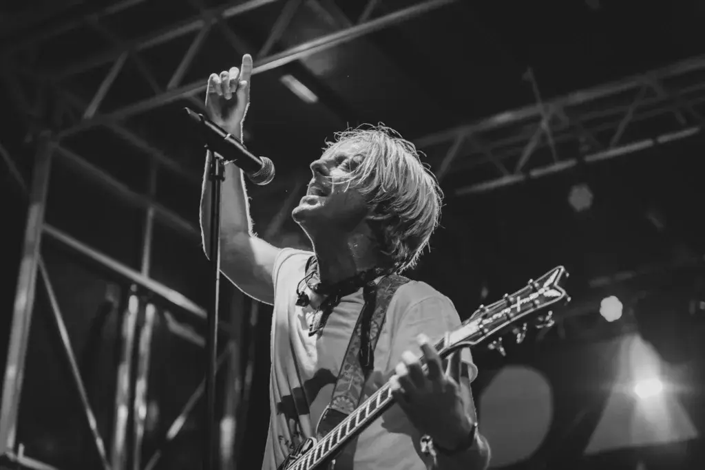 A performer with blonde hair sings into a microphone while playing guitar on stage in black and white.