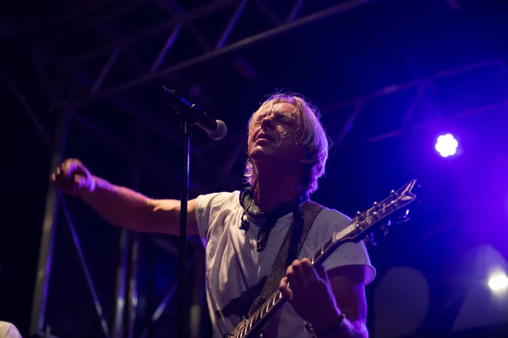 Performer singing and playing guitar on stage under purple stage lighting at an outdoor music festival.