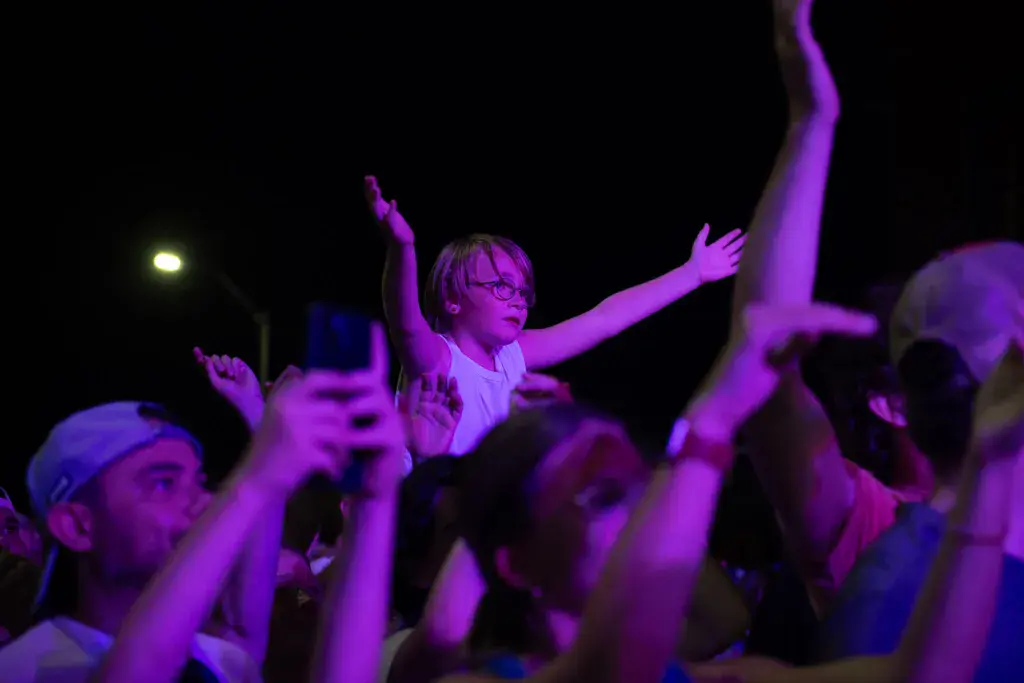 Festivalgoer crowd illuminated in purple light, with raised arms and hands holding phones during a nighttime performance.