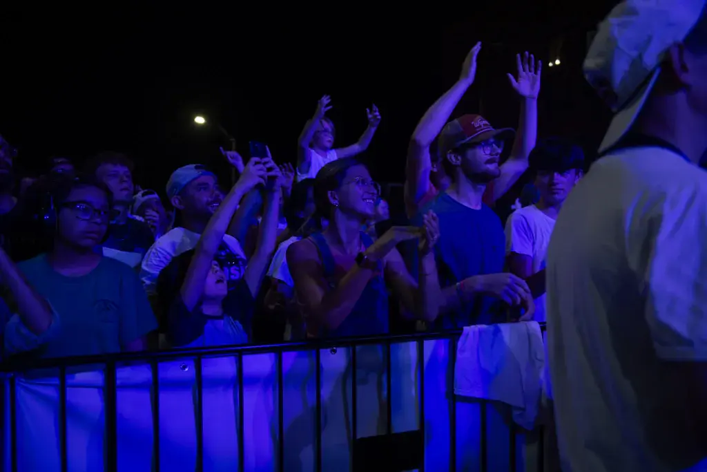 Enthusiastic crowd at night with raised hands and arms behind a barrier, bathed in blue stage lighting.