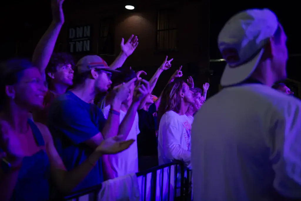 Enthusiastic crowd members raise their hands and cheer at an indoor concert venue bathed in purple stage lighting.