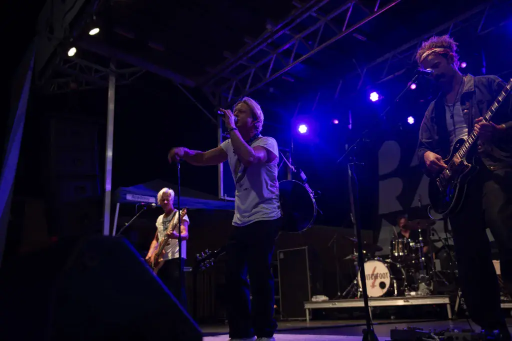A band performs on stage under blue lighting at an outdoor music festival, with multiple musicians playing instruments.