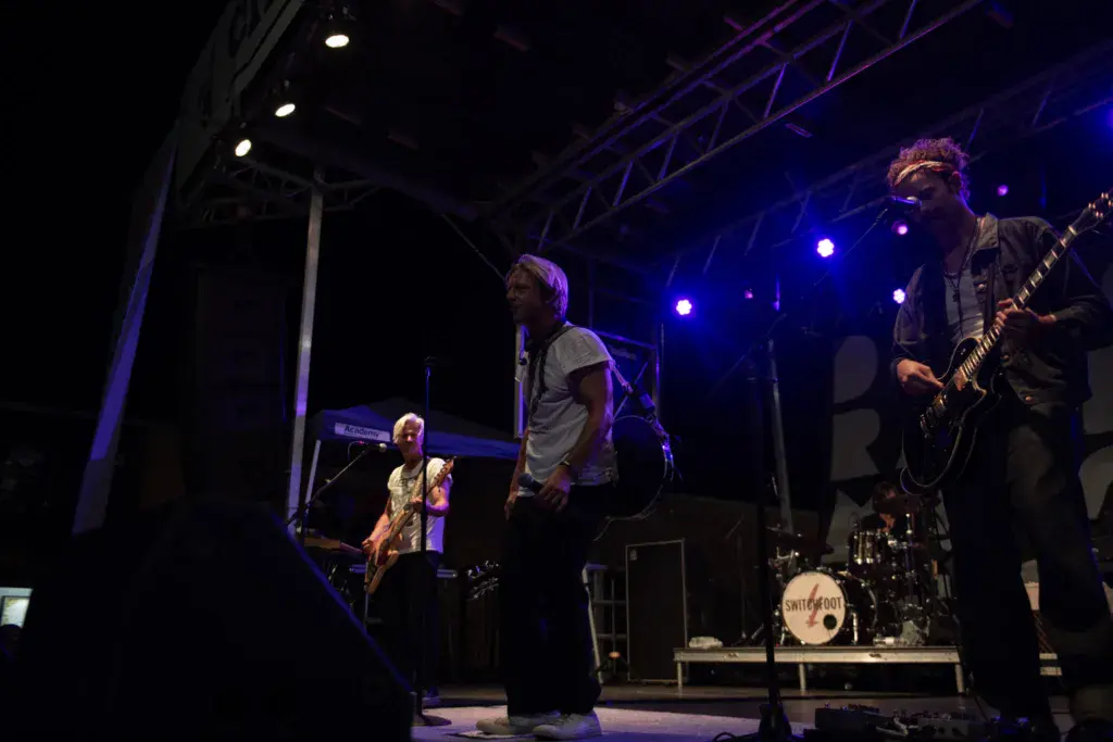 A four-piece band performs on an illuminated stage with blue lighting during an evening concert.