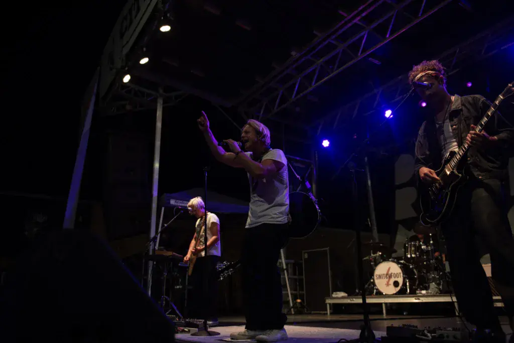 A band performs on stage at night with blue lighting, featuring a vocalist, guitarist, and drummer.