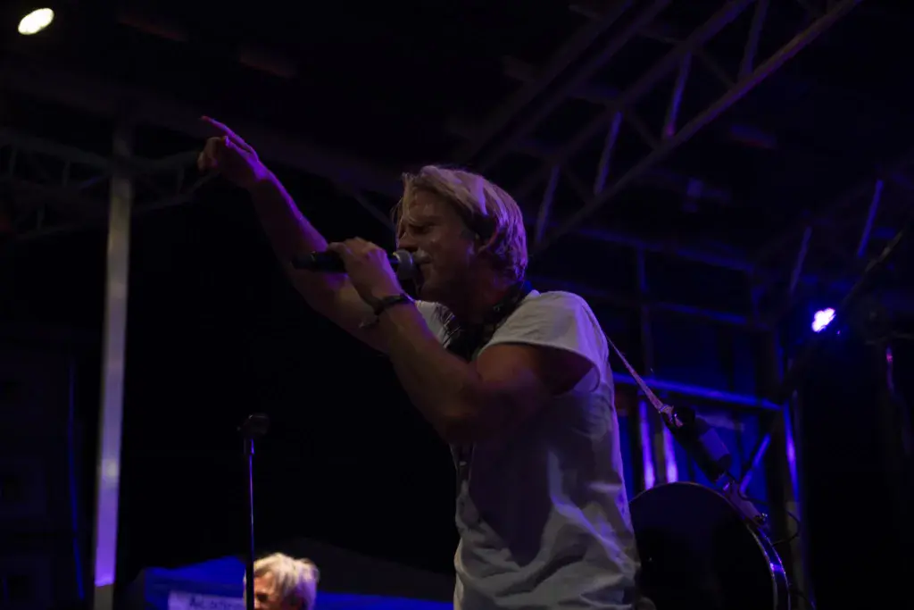 A performer on stage at night, singing into a microphone with arm raised, bathed in purple stage lighting.
