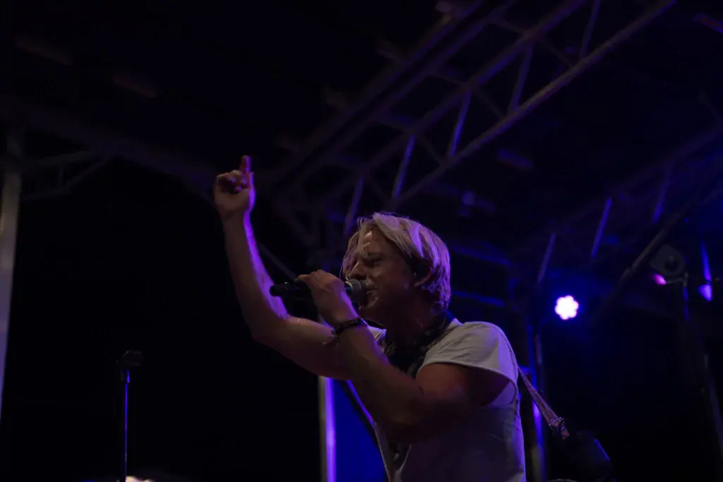 A performer on stage under purple lighting, singing into a microphone with one arm raised.