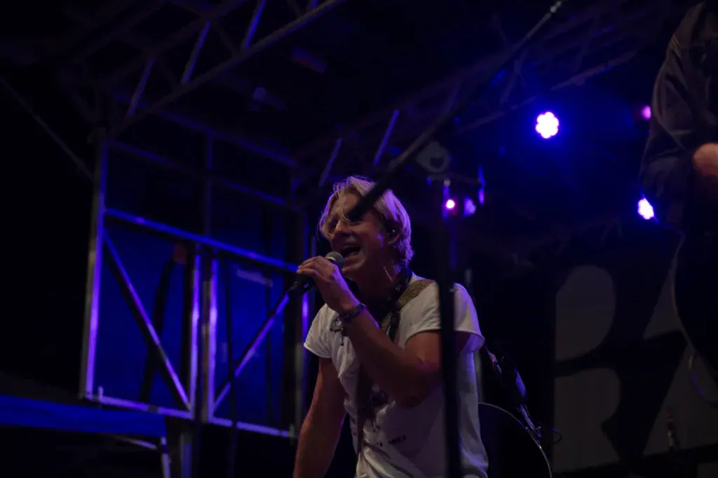 A performer sings into a microphone on stage under blue lighting at an outdoor music festival.