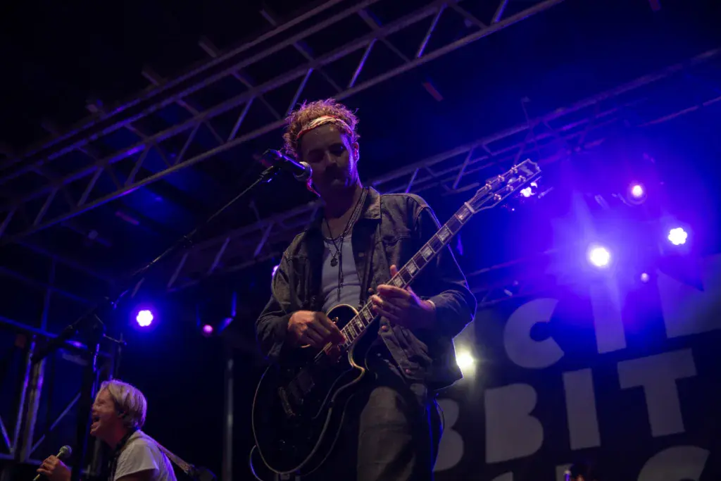 Guitarist performs on stage under blue lighting at an outdoor music festival.