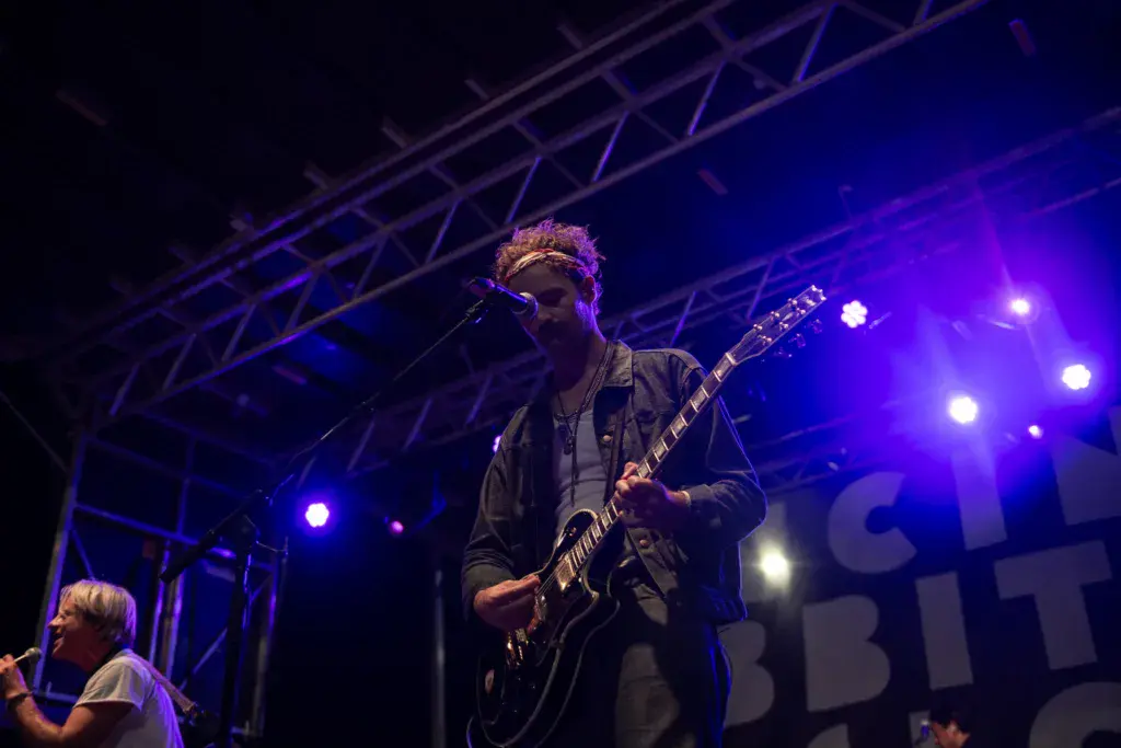 A guitarist performs on stage under purple and blue stage lights during an evening performance.