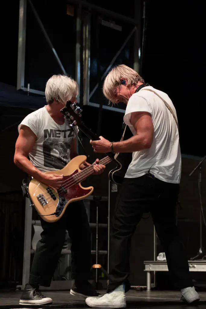 Two musicians perform on stage at Dancing Rabbit Music Festival, one singing at a microphone and one playing bass guitar.