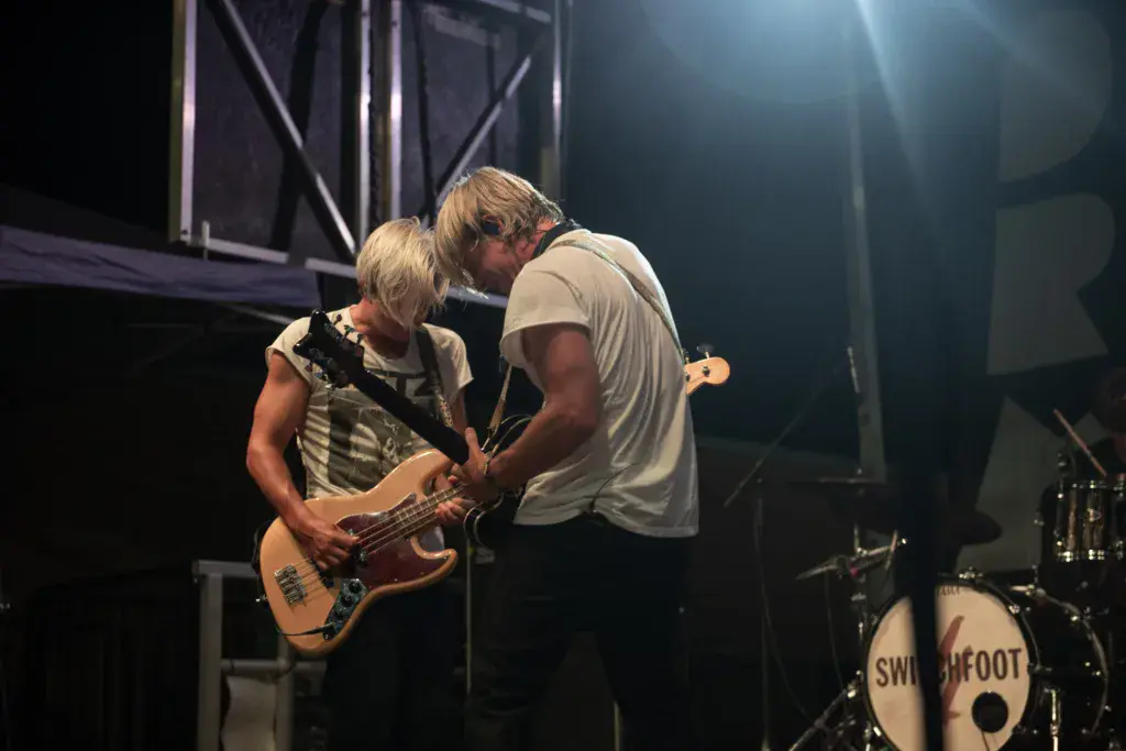 Two musicians perform on stage at night, one playing bass guitar and one singing, with drum kit visible.