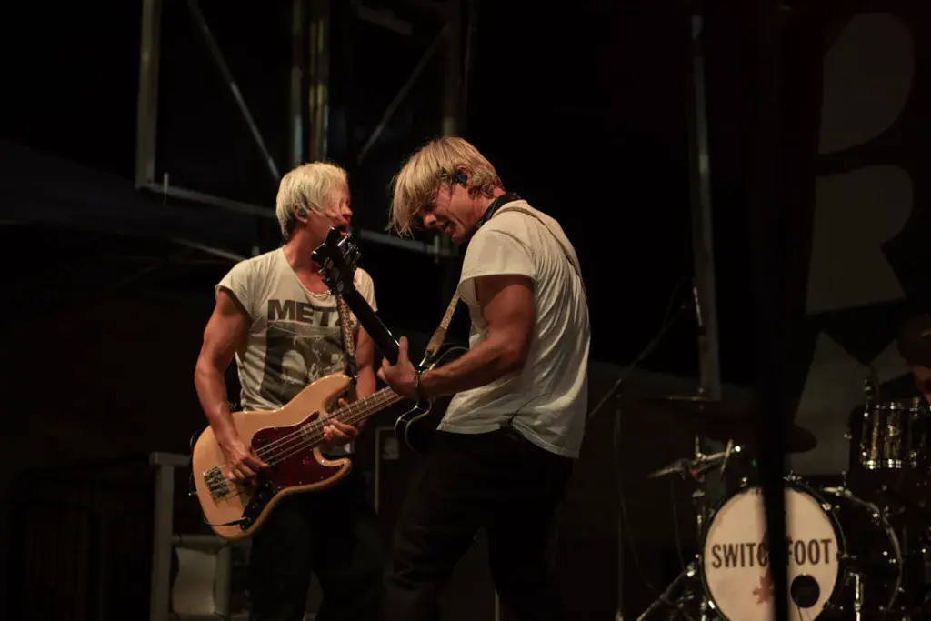 Two musicians perform on stage during Dancing Rabbit Music Festival, one singing into a microphone while playing bass guitar, the other singing.