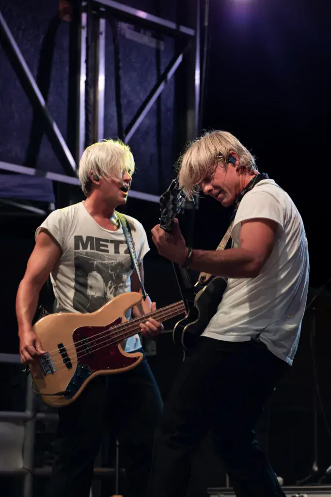 Two musicians perform on stage, one singing at a microphone while the other plays bass guitar.
