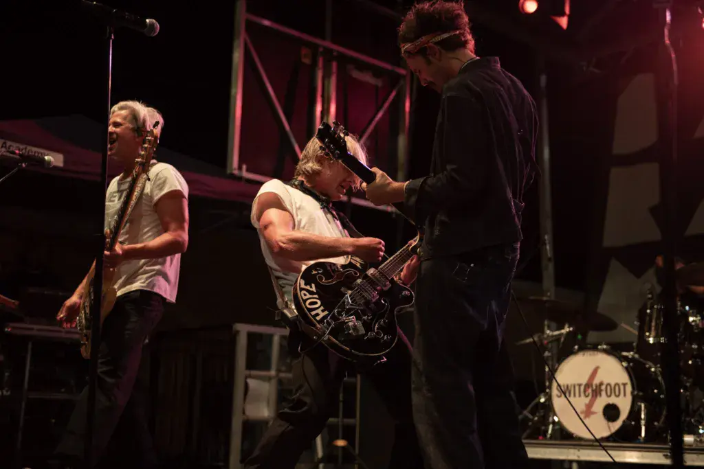 Three band members perform on stage under purple lighting, with a guitarist, bassist, and drummer visible.