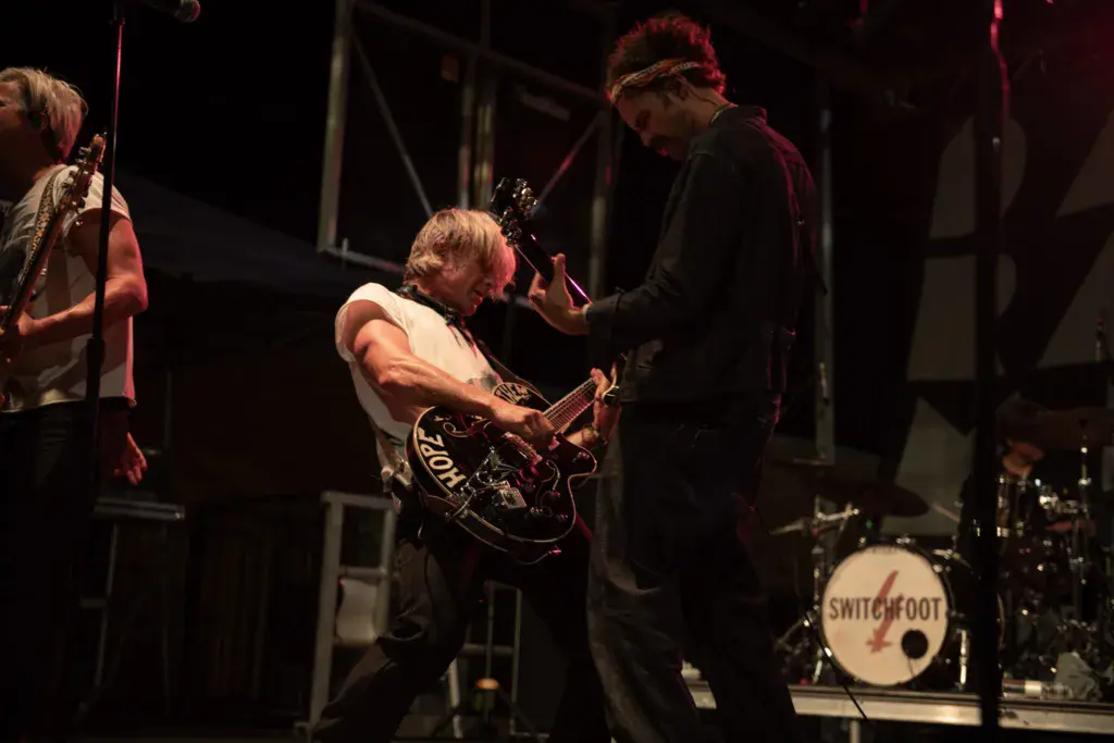 Band members perform on stage with guitar and drums during Dancing Rabbit Music Festival at night.