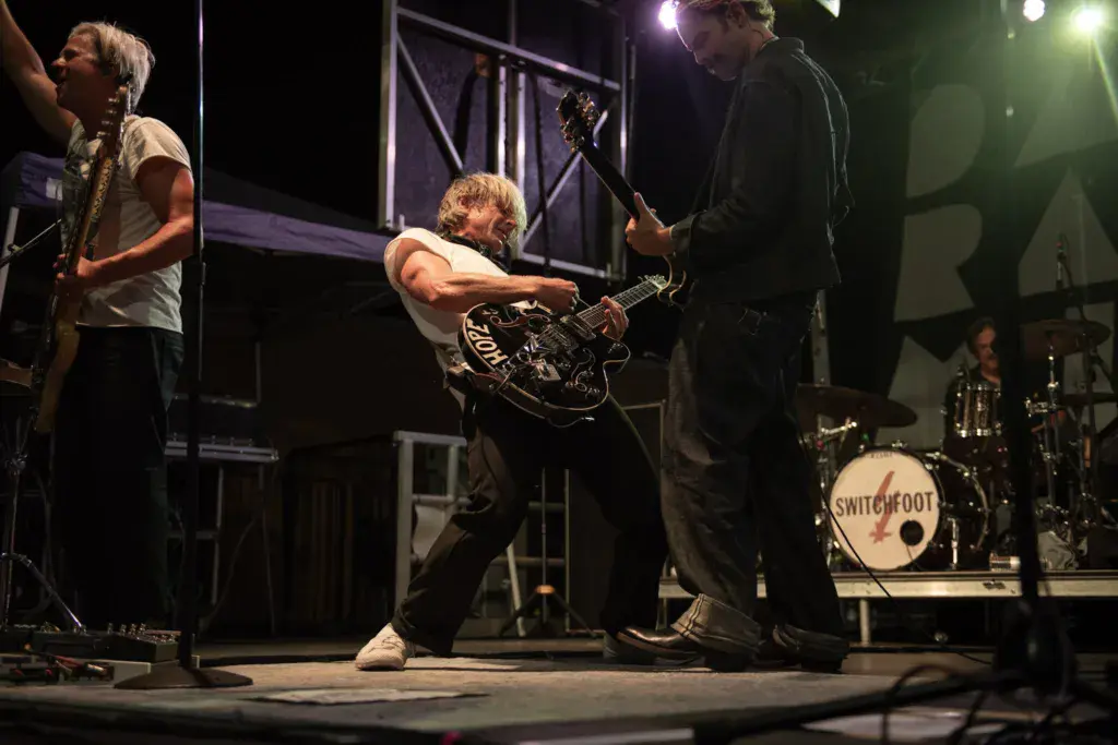 A rock band performs on stage at night, with guitarists and drummer visible under professional lighting.